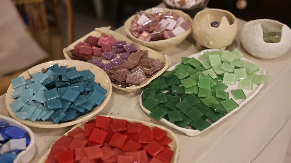 Bandejas con teselas de pasta vítrea en tonos verdes, azules, rojos y rosados sobre una mesa de taller de mosaico